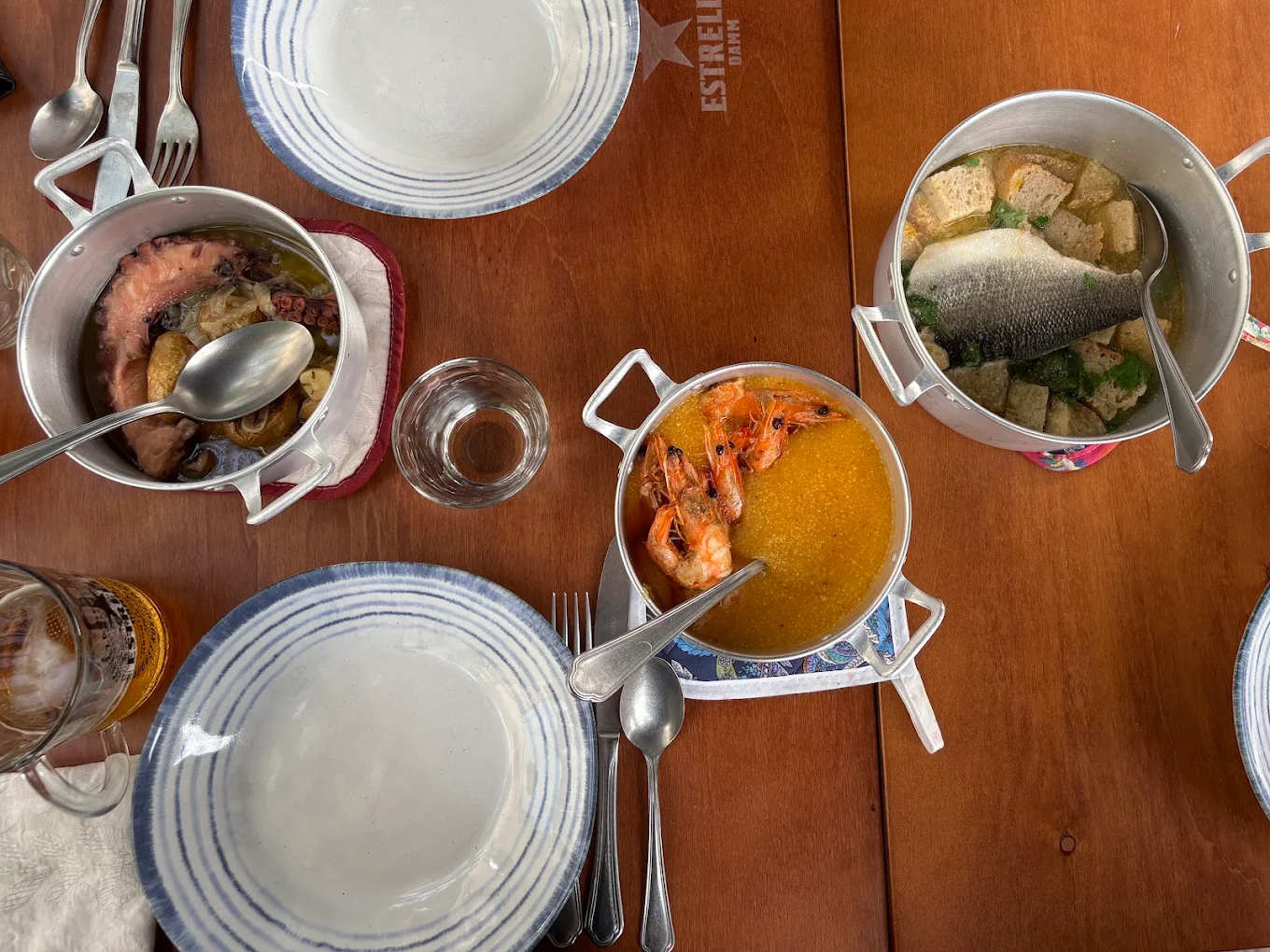 Mesa vista de cima com polvo, açorda de camarão e um peixe em caldo de pão, servidos em panelas para partilhar